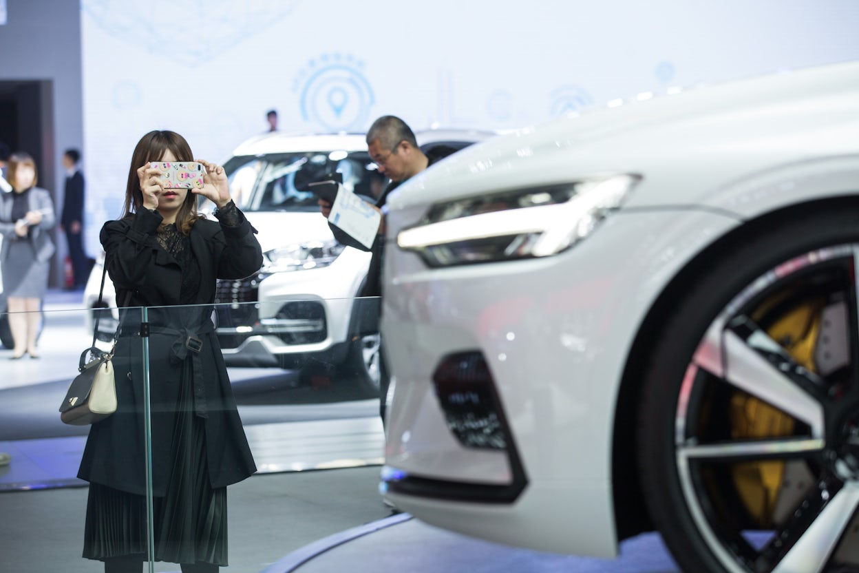 A woman photographing a white Polestar with her iPhone at car event.