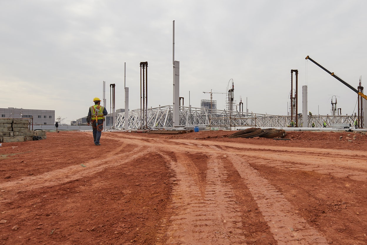 A worker wearing a safety helmet walking on red sand on a construction site.