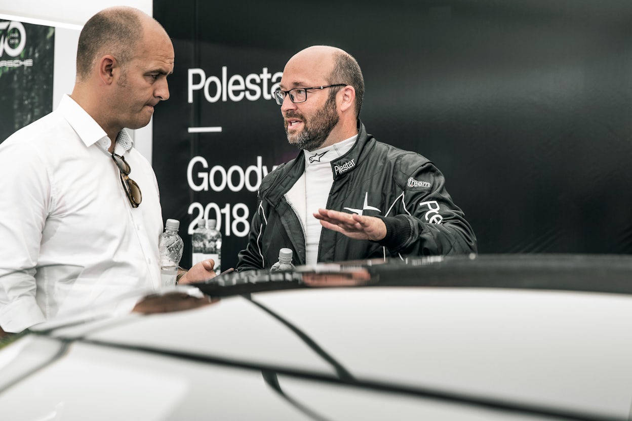 A man wearing a Polestar racing suit speaking to another man in a white shirt.