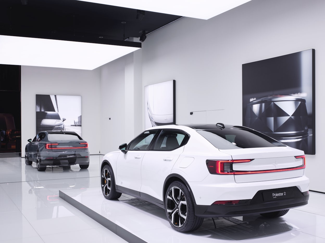 A white Polestar 2 on a stand with a grey Polestar in front of it on glossy white floor