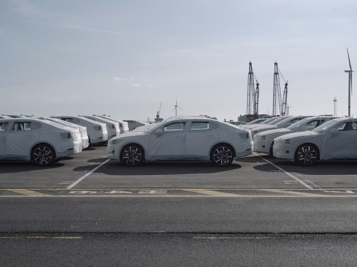Multiple Polestar cars covered with protective wrapping parked in rows at an outdoor lot, with cranes and wind turbines in the background.