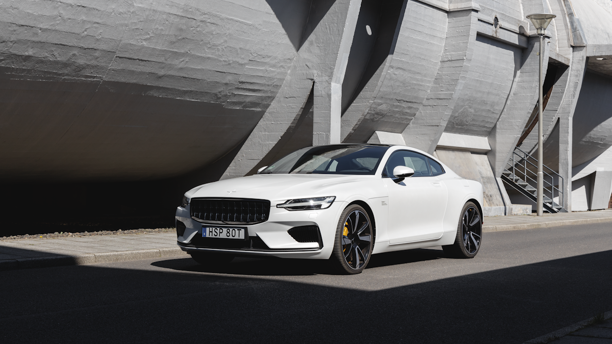 A white Polestar 1 in front of a curved concrete building.