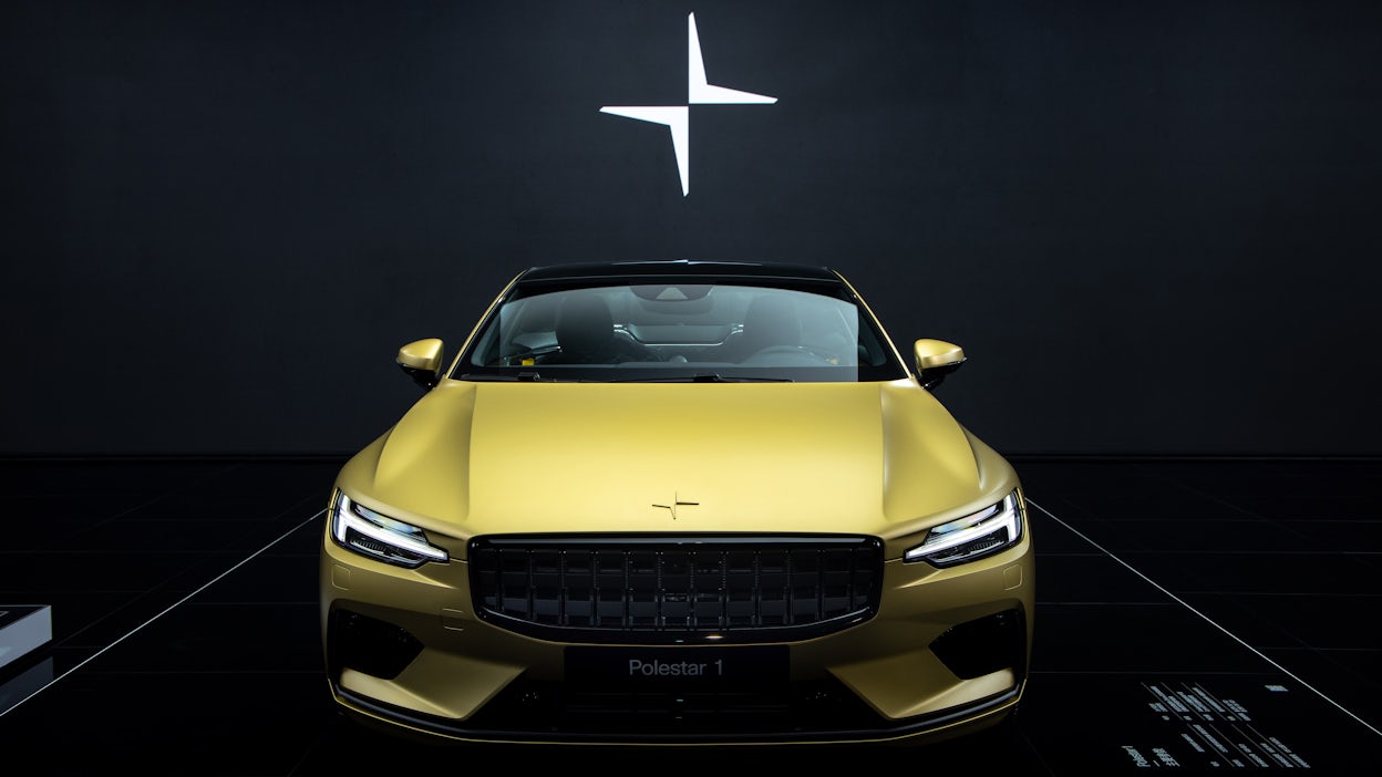 Front view of the Golden Polestar 1 in a dark room with an illuminated Polestar logo on the wall.