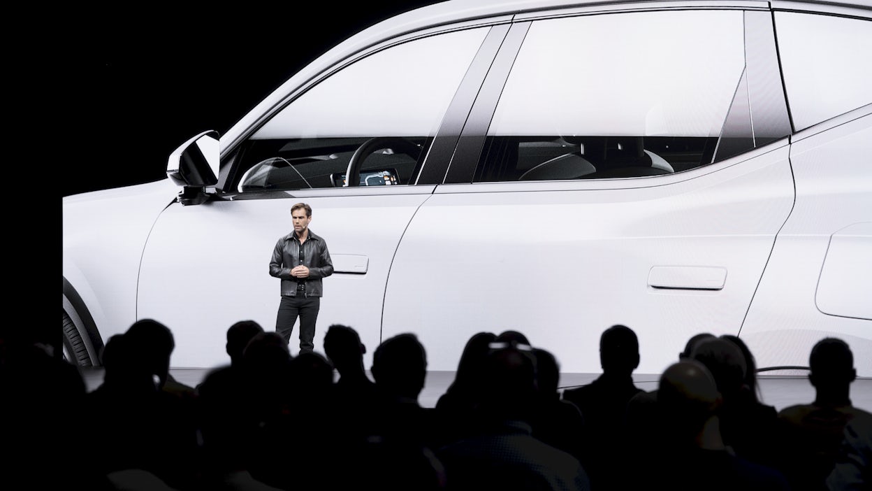 A man standing on stage in front of a screen showing a white car.