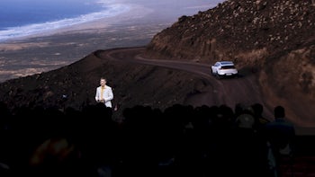 A man standing on stage in front of a screen showing a white car travelling down a mountiain road.