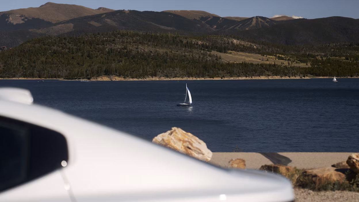 Polestar car in foreground, lake with sailboat and forested hills in background.