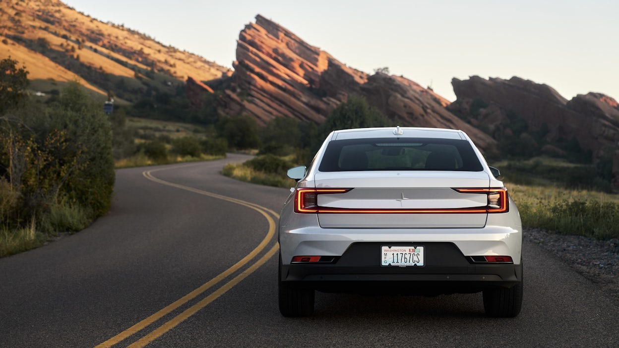 Back view of a white Polestar 2 driving on the road surrounded by a lush and rocky landscape.