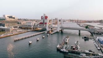 Cityscape with bridge, waterfront, boats, modern buildings, and red-and-white structure in the background.