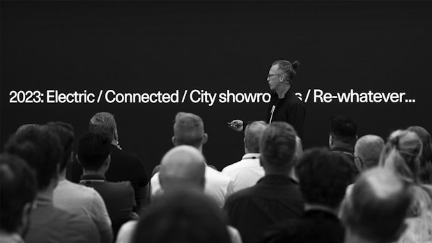 A person with a topknot hairstyle stands on a stage in front of a seated audience, presenting a slide with the text "2023: Electric / Connected / City showroom / Re-whatever...