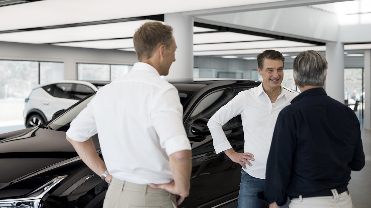 Three people standing near a black Polestar car in a showroom with another white Polestar car visible in the background.