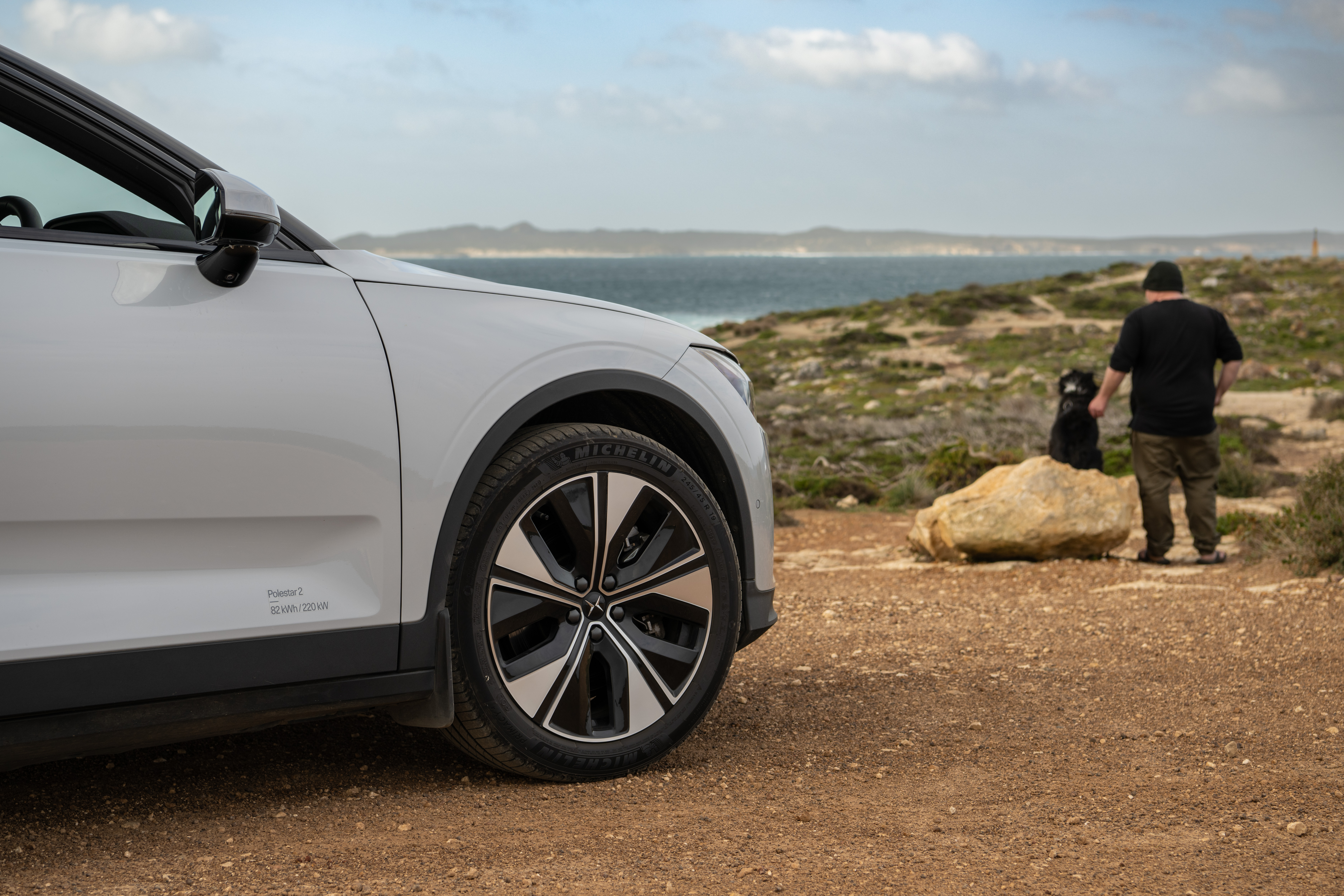 Polestar car parked on dirt road near coast, man and dog standing by rock in background.