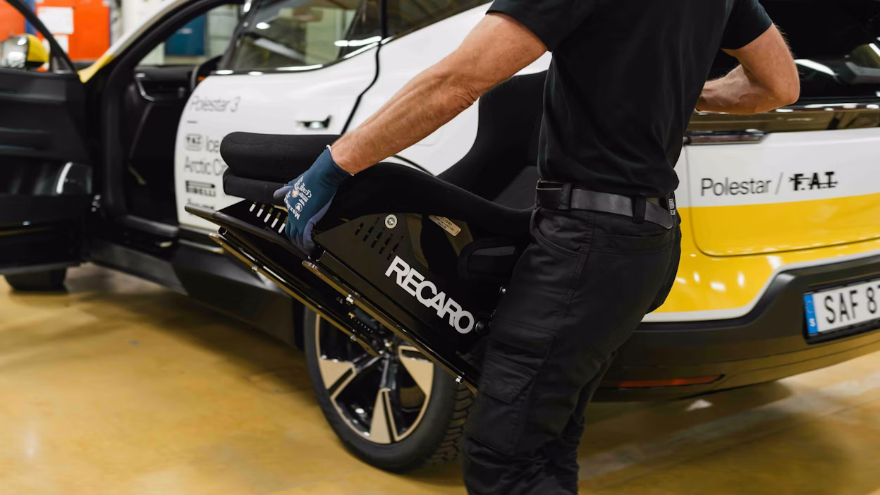 An engineer carrying a racing seat in front of a car
