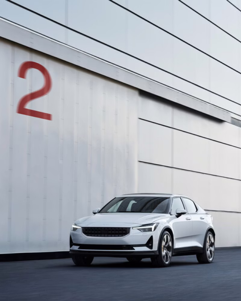 A white Polestar 2 parked infront of a grey garage with a big red 2 painted on the wall