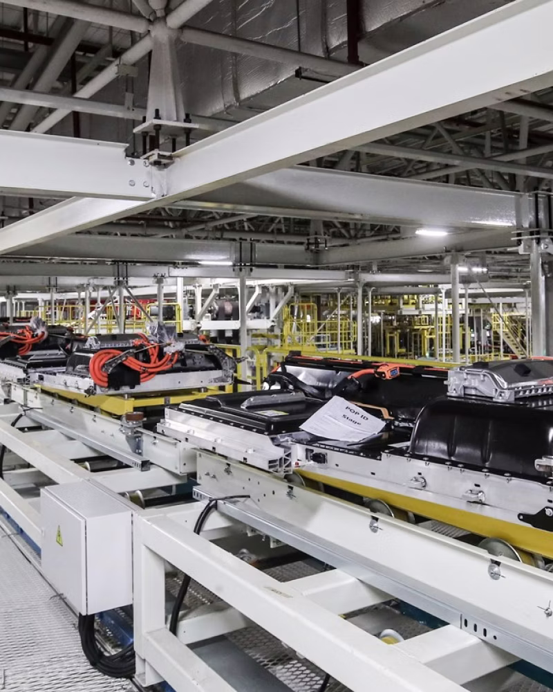 Battery packs on an assembly line in a factory with industrial equipment and structural beams visible.