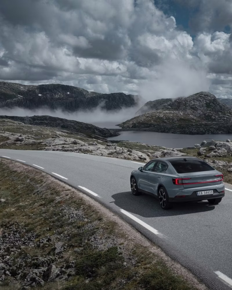 Polestar car driving on a curved road surrounded by rocky terrain and a lake under cloudy sky.