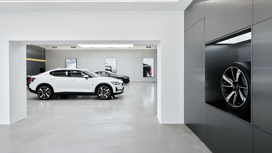 White Polestar 2 car in showroom with another car in background and a wheel displayed in a wall compartment.