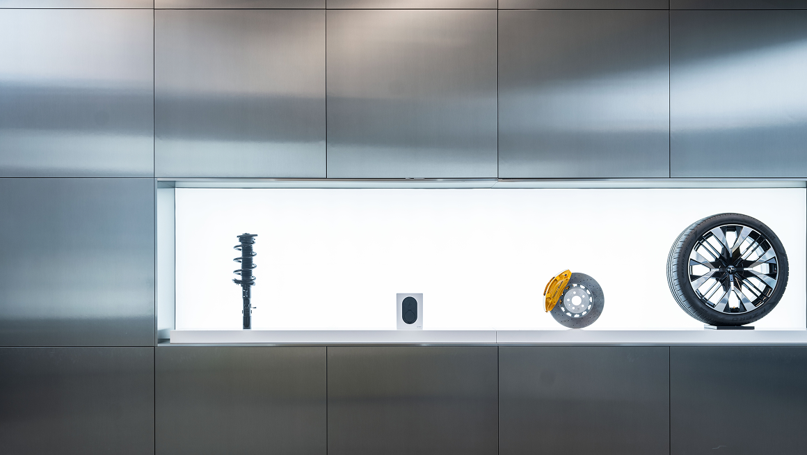 Display of car suspension, speaker, brake disc with caliper, and wheel against a metallic wall.