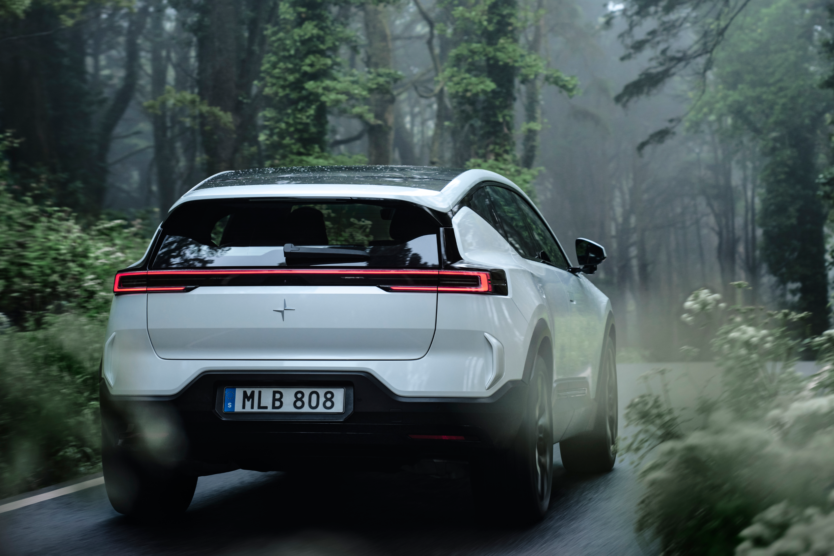 A rear view of a white Polestar 3 driving in a foggy environment with trees and green foliage.