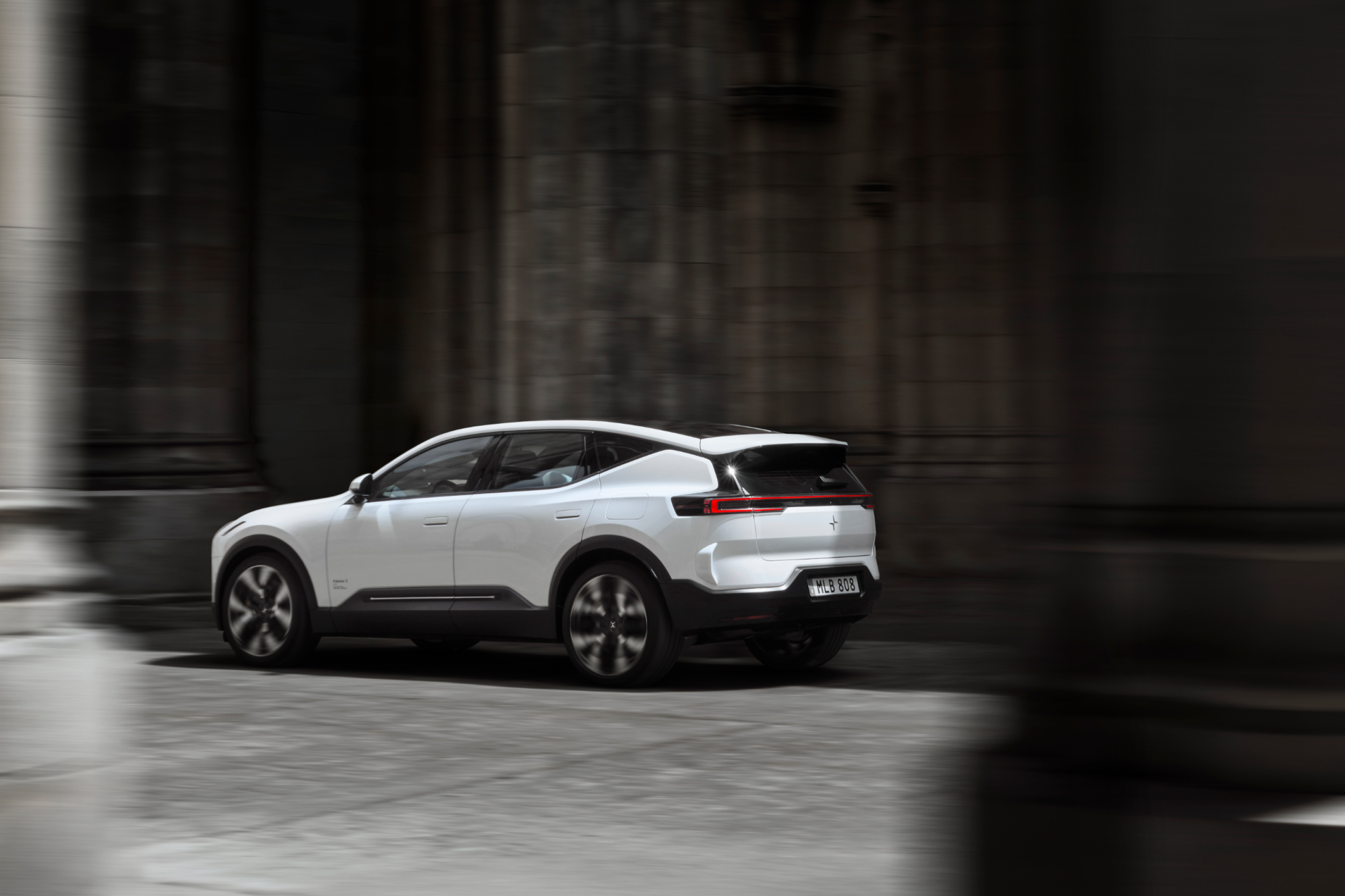 A rear-angled view of a white Polestar 3 driving between grey stone walls and columns.