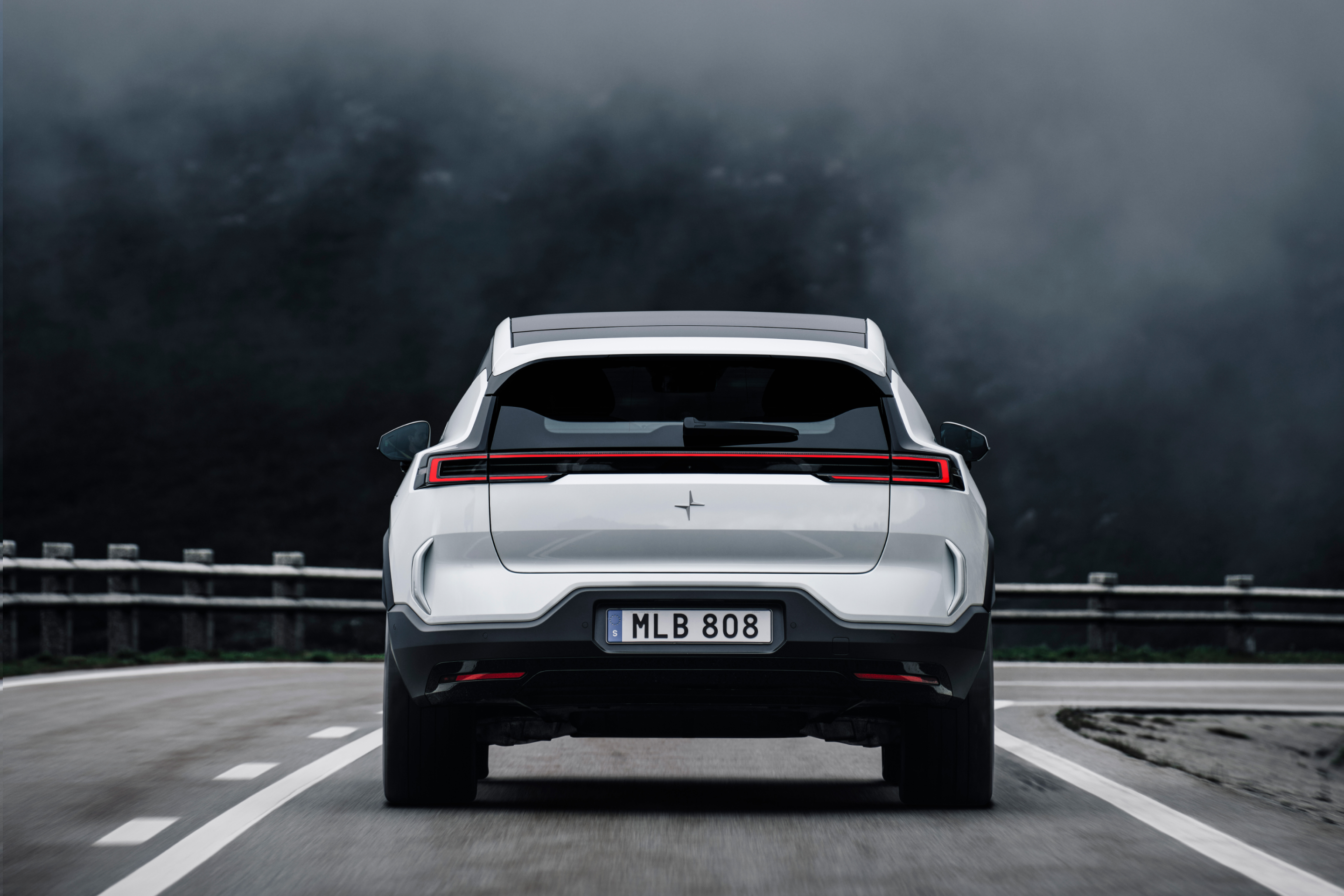 View of a white Polestar 3 from behind driving on a motorway against a foggy, mountainous backdrop.